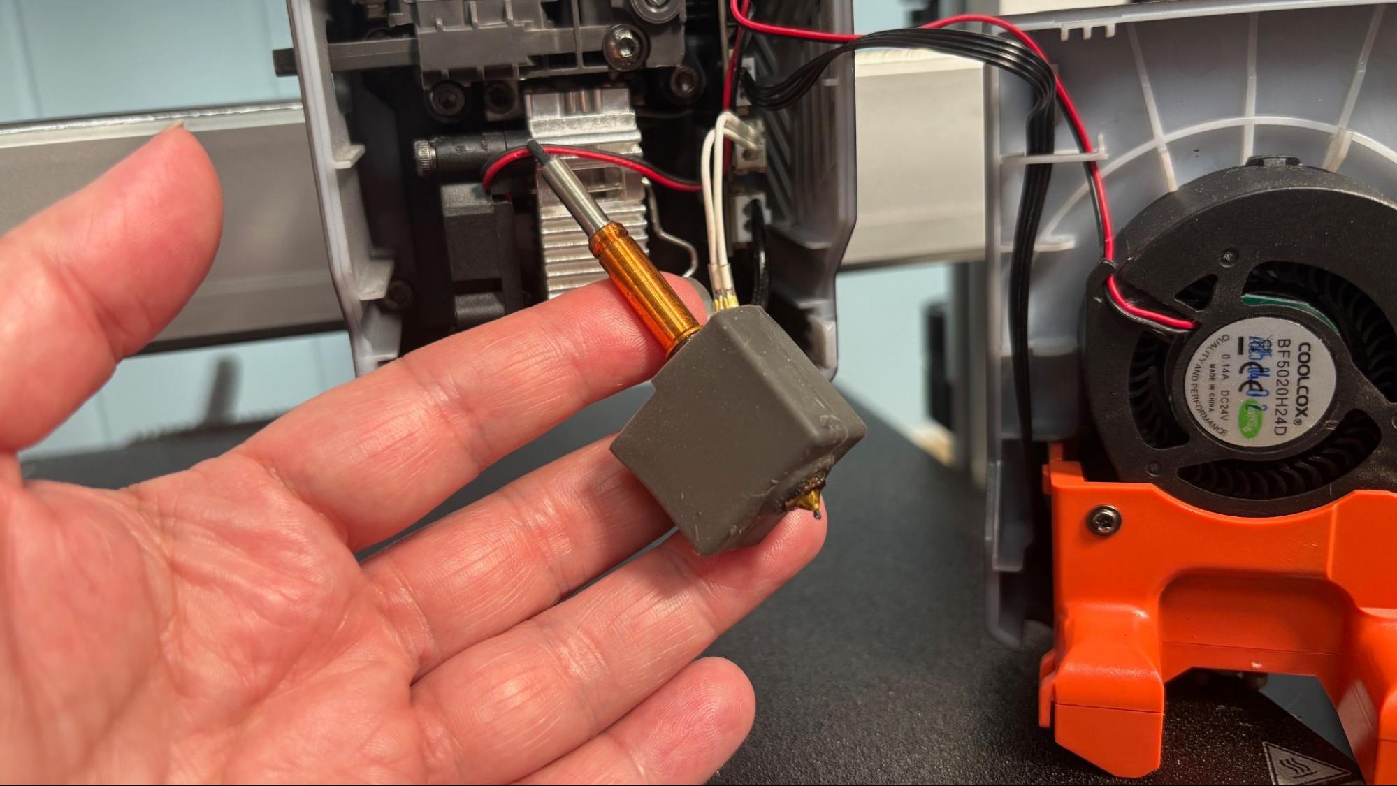 Снятый быстросъёмный хотенд Anycubic Kobra 3 V2 в руке, рядом открытая печатающая голова с вентилятором обдува